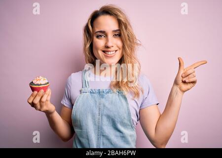 Junge schöne blonde Frau Eatimg Schokoladenkuchen über isoliertem rosafarbenem Hintergrund sehr glücklich, mit Hand und Finger zur Seite zu zeigen Stockfoto