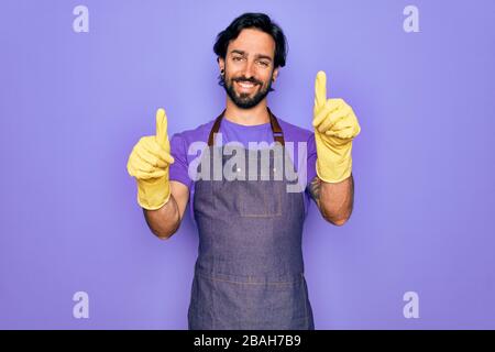 Junger gutaussehender hispanischer Klenaer, der eine Schürze für Hausarbeit und Waschhandschuhe trägt und positive Geste mit der Hand abgibt, mit dem Daumen lächelnd und glücklich Stockfoto