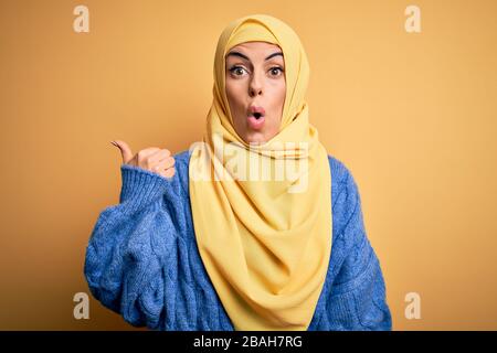 Junge, schöne brünette muslimin, die arabischen Hijab über isoliertem gelbem Hintergrund trägt, überraschte mit dem Handfinger zur Seite, offenen Mund a Stockfoto
