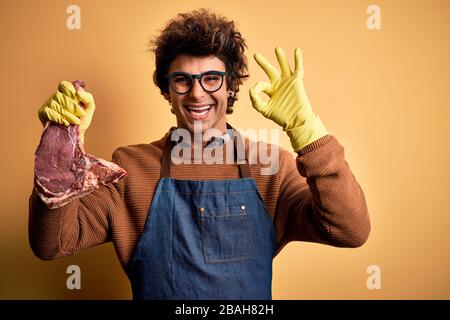 Junger gutaussehender Metzgermeister, der Steak hält, der über isoliertem gelbem Hintergrund steht, lächelnd positiv, wobei er mit Hand und Fingern ein gutes Zeichen tat. Erfolgreich Stockfoto