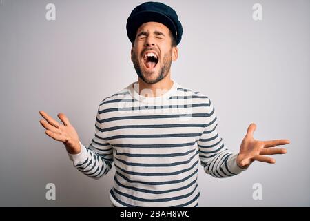 Junger gutaussehender Seemannsmann mit Bart, marineblaue gestreifte Uniform und Kapitänshut verrückt und verrückt schreien und mit aggressivem Ausdruck und ar schreien Stockfoto