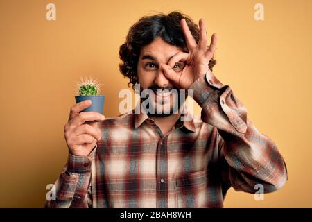 Junger Mann mit lockigem Haar und Bart, der den kleinen Kaktuspflanzentopf über gelbem Hintergrund hält, mit einem fröhlichen Gesicht, das lächelt, und einem guten Schild mit Hand auf Augenschmaus tut Stockfoto