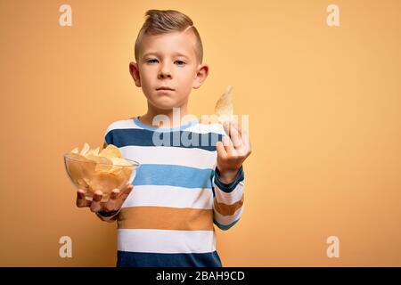 Junge kleine kaukasische Kinder, die nicht hitzelige Kartoffeln essen, knacken Pommes über gelben Hintergrund mit einem selbstbewussten Ausdruck auf einem klugen Gesicht, das ernsthaft denkt Stockfoto