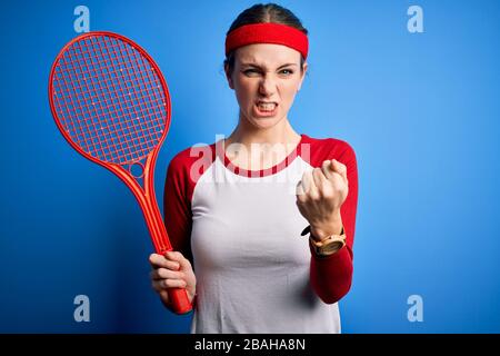 Junge schöne Rotkopfsportlerin, die mit Schläger über blauem Hintergrund Tennis spielt, ärgerte sich und frustrierte Schreie mit Wut, verrücktem und schreiendem Witz Stockfoto