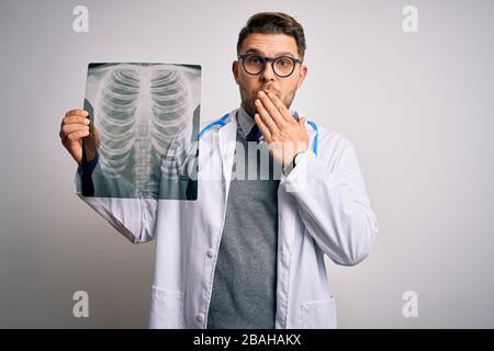 Junger Arzt, der einen medizinischen Mantel trägt, der auf die Röntgenaufnahme des Brustkorbs über dem isolierten Mund der Hintergrundabdeckung blickt, mit der Hand, die aus versehen mit Scham schockiert ist, Stockfoto