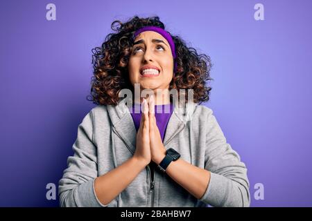 Schöne, lockige arabische Sportlerin, die Sport macht und Sportswear über purpurem Hintergrund trägt, bettelt und betet mit den Händen zusammen mit dem Hope Expression On Stockfoto