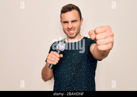 Junger gutaussehender Sänger mit blauen Augen, der mit Mikrofon über weißem Hintergrund singt, ärgerte sich und frustrierte Schreie mit Wut, Wahnsinn und Geschrei Stockfoto