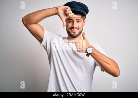 Junger Fahrer mit Bart und Hut, der über einem isolierten weißen Hintergrund steht und lächelt, um mit den Händen und Fingern ein fröhliches Gesicht zu bilden. Creativit Stockfoto