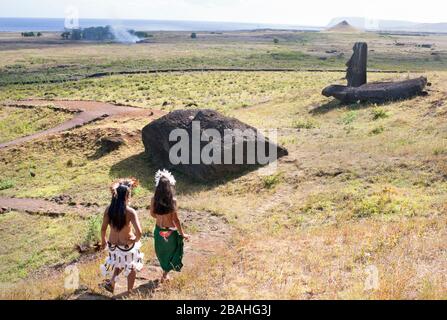 Frauen von Rapa Nui und Tänzer, die auf dem Weg am gefallenen Moai im Rano Raraku Moai-Steinbruch auf der Osterinsel vorbeigehen Stockfoto