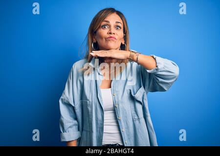 Schöne Frau im mittleren Alter, die Casual Hemd trägt, das über isoliertem blauem Hintergrund steht und mit der Hand als Messer die Kehle durchschneidet, bedroht die Aggression mit Pelz Stockfoto