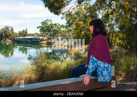 Ein einziger weiblicher Tourist mit einer roten Schal sitzt auf einer Gartenbank, die über den Teich auf der Pheasant Farm im Barossa-Tal blickt. Stockfoto