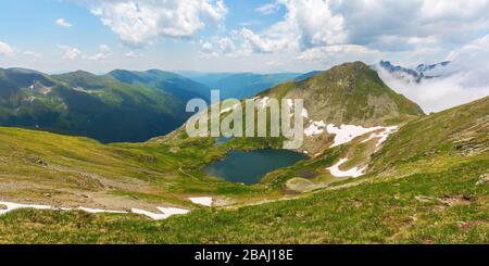 Schöne Natur der rumänischen Berge. capra-see im Tal. Von Gras, Felsen und Schnee bedeckte Hügel. Wunderbarer sonniger Sommertag mit wunderbarer Clou Stockfoto