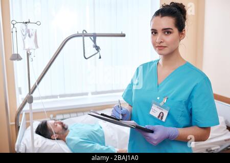 Portrait der ernsthaften jungen Krankenschwester in den chirurgischen Handschuhen, die Hinweise zur Gesundheit der Patienten macht, während sie die medizinische Karte im Krankenhaus füllt Stockfoto