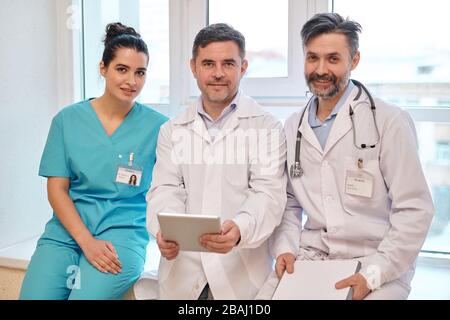 Team von erfahrenen medizinischen Mitarbeitern im einheitlichen Sitzen auf Fensterbank im Krankenhaus und Blick auf die Kamera Stockfoto