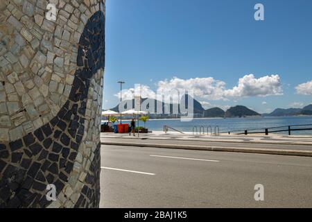 Dekorative Wand mit portugiesischen Fliesenwellen mit dem Berg Sugarloaf hinter einer leeren Straße, einem Boulevard und einem Strand in Copacabana Stockfoto