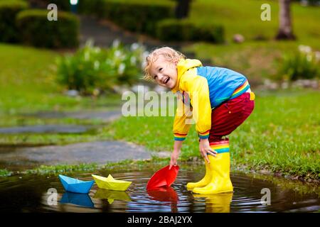 Kinder spielen mit Papier Boot in der Pfütze. Kinder spielen im Freien im Herbst regen. Fallen Regen im Freien Aktivitäten für junge Kinder. Kid in Schlamm springen Stockfoto