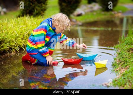 Kinder spielen mit Papier Boot in der Pfütze. Kinder spielen im Freien im Herbst regen. Fallen Regen im Freien Aktivitäten für junge Kinder. Kid in Schlamm springen Stockfoto