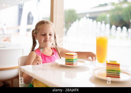 Kinder essen Regenbogenkuchen im Restaurant. Kleines Mädchen mit Süßigkeiten und Gebäck. Kind trinkt frischen Orangensaft im Café. Familie, die in der Cafeteri der Stadt auswärts isst Stockfoto