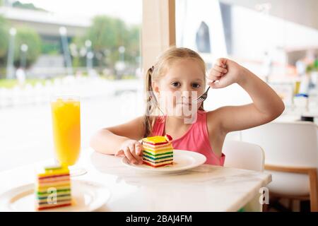 Kinder essen Regenbogenkuchen im Restaurant. Kleines Mädchen mit Süßigkeiten und Gebäck. Kind trinkt frischen Orangensaft im Café. Familie, die in der Cafeteri der Stadt auswärts isst Stockfoto