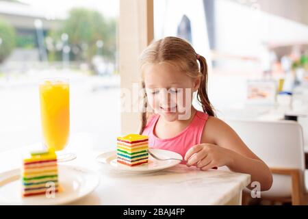 Kinder essen Regenbogenkuchen im Restaurant. Kleines Mädchen mit Süßigkeiten und Gebäck. Kind trinkt frischen Orangensaft im Café. Familie, die in der Cafeteri der Stadt auswärts isst Stockfoto