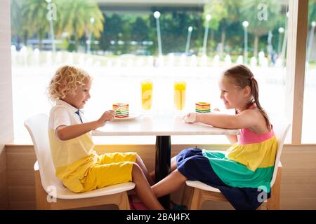 Kinder essen Regenbogenkuchen im Restaurant. Junge und Mädchen mit Süßigkeiten und Gebäck. Kinder trinken frischen Orangensaft im Café. Familie, die im Stadtcafé auswärts isst Stockfoto