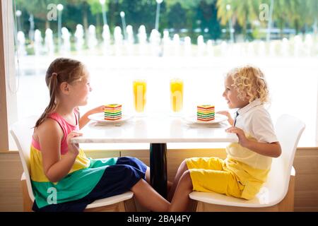 Kinder essen Regenbogenkuchen im Restaurant. Junge und Mädchen mit Süßigkeiten und Gebäck. Kinder trinken frischen Orangensaft im Café. Familie, die im Stadtcafé auswärts isst Stockfoto