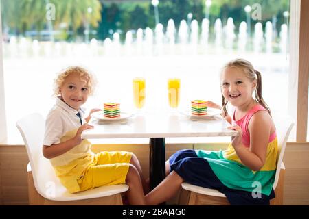 Kinder essen Regenbogenkuchen im Restaurant. Junge und Mädchen mit Süßigkeiten und Gebäck. Kinder trinken frischen Orangensaft im Café. Familie, die im Stadtcafé auswärts isst Stockfoto