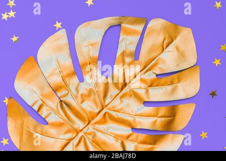 Goldbemaltes Blatt aus Monstera und sternförmigen Konfetti auf violettem Hintergrund. Modisches Layout für Design Stockfoto