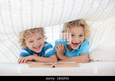 Lesebuch für Kinder im Bett unter der Strickdecke. Zwei Brüder spielen zusammen. Kinder gemütliches Zimmer im Hyggge-Stil. Kleiner Junge, der Hausaufgaben macht, bevor er schläft. Stockfoto