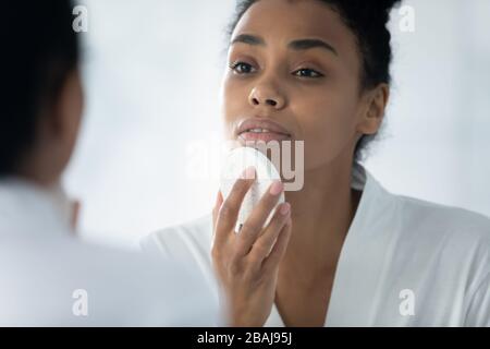 Afrikanerin, die im Spiegel reflektiert und den Gesichtsschwamm hält, reinigt das Gesicht Stockfoto