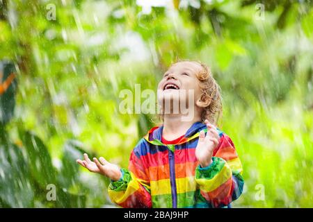 Kind spielt im Regen am sonnigen Herbsttag. Kinder unter schwerer Dusche mit Regenbogenjacke. Kleiner Junge im regnerischen Park. Outdoor-Aktivitäten im Herbst Stockfoto