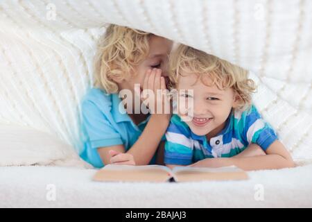 Lesebuch für Kinder im Bett unter der Strickdecke. Zwei Brüder spielen zusammen. Kinder gemütliches Zimmer im Hyggge-Stil. Kleiner Junge, der Hausaufgaben macht, bevor er schläft. Stockfoto