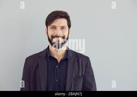 Positiver bärtiger Kerl in einer Jacke lächelnd sieht die Kamera auf grauem Hintergrund an. Stockfoto