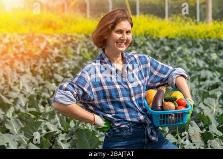 Frau, die nach der Arbeit auf dem Feld ruht und einen Korb mit geerntetem Gemüse hält. Frau, die an einem sonnigen Tag auf einem landwirtschaftlichen Feld arbeitet Stockfoto