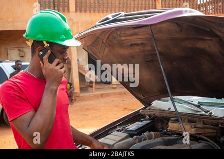 Ein junger afrikanischer Ingenieur, der telefoniert, während er an einem Auto eines Kunden arbeitet. Stockfoto