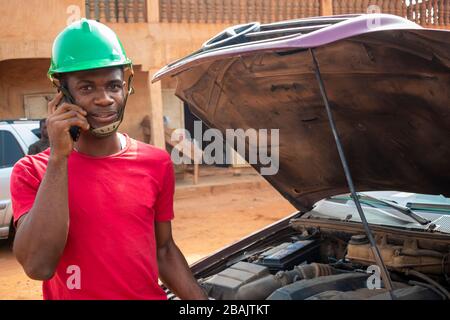 Ein junger afrikanischer Ingenieur, der telefoniert, während er an einem Auto eines Kunden arbeitet. Stockfoto