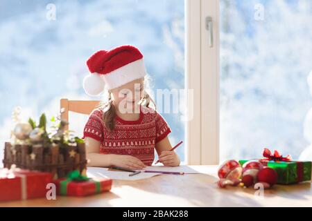 Kind schreiben Brief an Santa am Heiligabend. Kinder schreiben Weihnachtsgeschenk Wunschliste. Kleines Mädchen sitzen in eingerichtetes Wohnzimmer mit grossen Fenster auf Sun Stockfoto