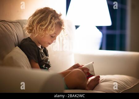 Lesebuch für Kinder. Kinder lesen Bücher. Kleiner Junge, der auf der weißen Couch im dunklen Wohnzimmer sitzt und Bilder im Buch sieht. Kind macht Hausaufgaben für ELE Stockfoto