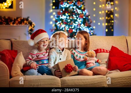 Kinder am Weihnachtsbaum und Kamin am Weihnachtsabend. Familie mit Kindern, die Weihnachten zu Hause feiern. Junge und Mädchen in Strickpullover auf weißer Couch