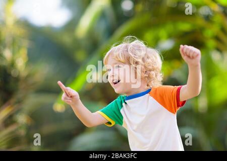 Kinder, die im sonnigen Sommerpark spielen. Kinder laufen im Freien. Kleiner blonder lockiger Junge, der lustige Gesichter macht. Gartenspaß. Stockfoto