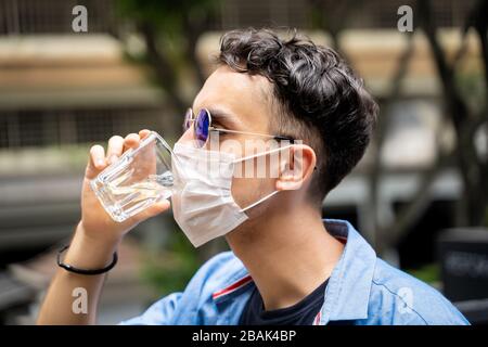 Profil eines jungen Mannes mit Sonnenbrille Trinkwasser, während er eine Schutzmaske auf seinem Gesicht trägt. Covid-19 Stockfoto