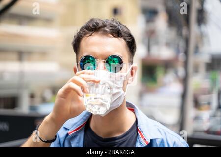 Junger Mann mit reflektierender Sonnenbrille, der Wasser trinkt, während er eine Schutzmaske auf seinem Gesicht trägt. Covid-19 Stockfoto