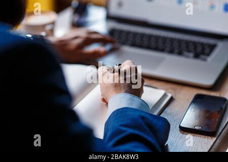Der Geschäftsmann im Büro macht Notizen in einem Notizbuch. Ein Student arbeitet in einem Laptop hinter einem Laptop. Nahaufnahme von Computer, Händen und Notebook Stockfoto