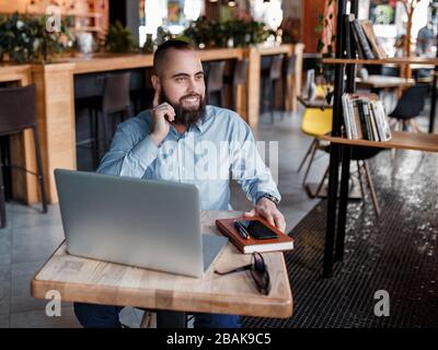 Der junge lächelnde bärtige Geschäftsmann sitzt am Tisch vor dem Computer, spricht auf dem Handy und hält Stift. Freiberufler arbeitet zu Hause. Telefon-Co Stockfoto