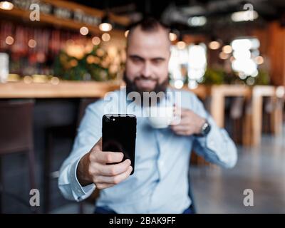 Am Morgen folgt der Geschäftsmann den Nachrichten, dem Aktienkurs per Telefon und trinkt Kaffee. Junger Investor. Bärtiger Mann mit einem Lächeln schaut in Stockfoto
