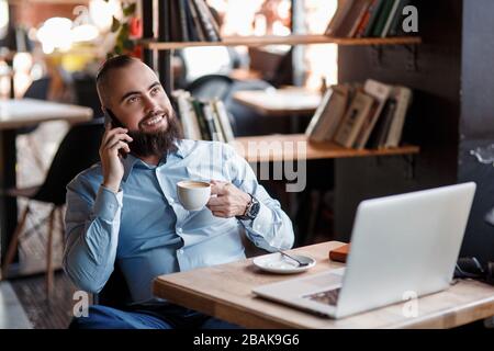 Der junge lächelnde bärtige Geschäftsmann sitzt am Tisch vor dem Computer, spricht auf dem Handy und hält Stift. Freiberufler arbeitet zu Hause. Telefon-Co Stockfoto