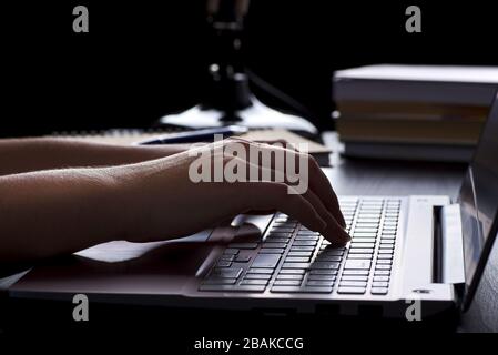 Stillleben von Arbeitsstunden oder Büro nachts unter Lampenleuchten. Junge Freiberuflerin tippt auf Laptop-Tastatur und macht Hausaufgaben. Stockfoto