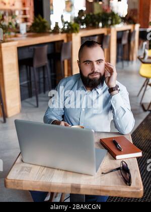 Der junge lächelnde bärtige Geschäftsmann sitzt am Tisch vor dem Computer, spricht auf dem Handy und hält Stift. Freiberufler arbeitet zu Hause. Telefon-Co Stockfoto