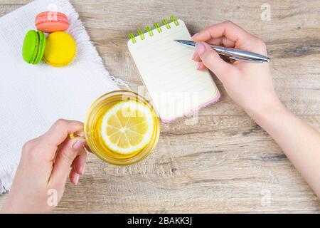 Die Hand hält einen silbernen Stift und schreibt in einem offenen Notizbuch, neben einem Notizbuch auf einem weißen Handtuch gibt es Minztee mit Zitronen- und mehrfarbigen Muffins Stockfoto