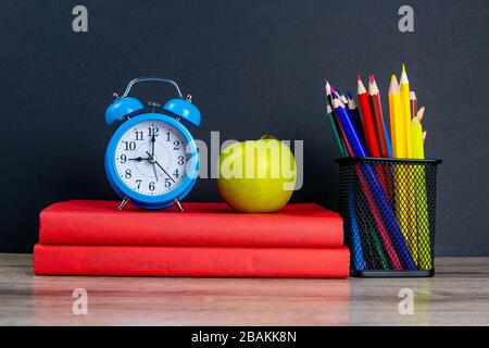 Der blaue Wecker sowie mehrfarbige Bleistifte stehen auf einem Stapel roter Bücher, neben einem apfel Stockfoto